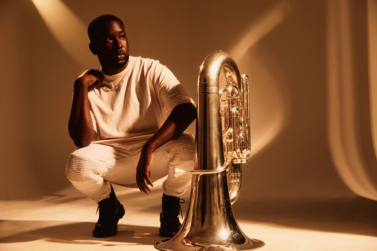 Tuba player Theon Cross is dressed in white, crouching beside a tube.