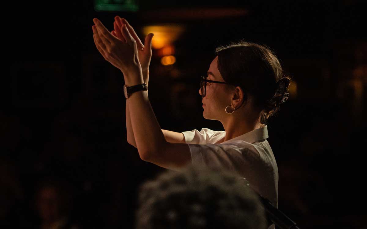 Olivia Murphy conducts musicians out of the shot, her arms are raised as if she is about to clap, Her hair is tied back in a bun and she wears glasses and a loose white shirt.