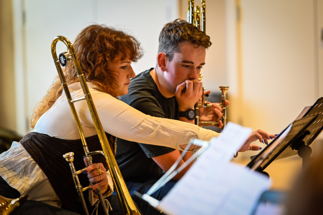 A female trombonist in her early 20s, pointing at a sheet of music to explain something to a teenage boy who is learning the trombone.