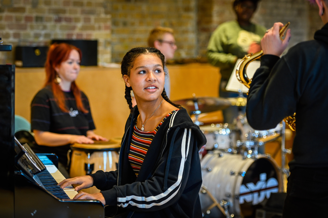 A teenage girl playing the piano, looking up at an educator on the saxophone who is explaining something to her.