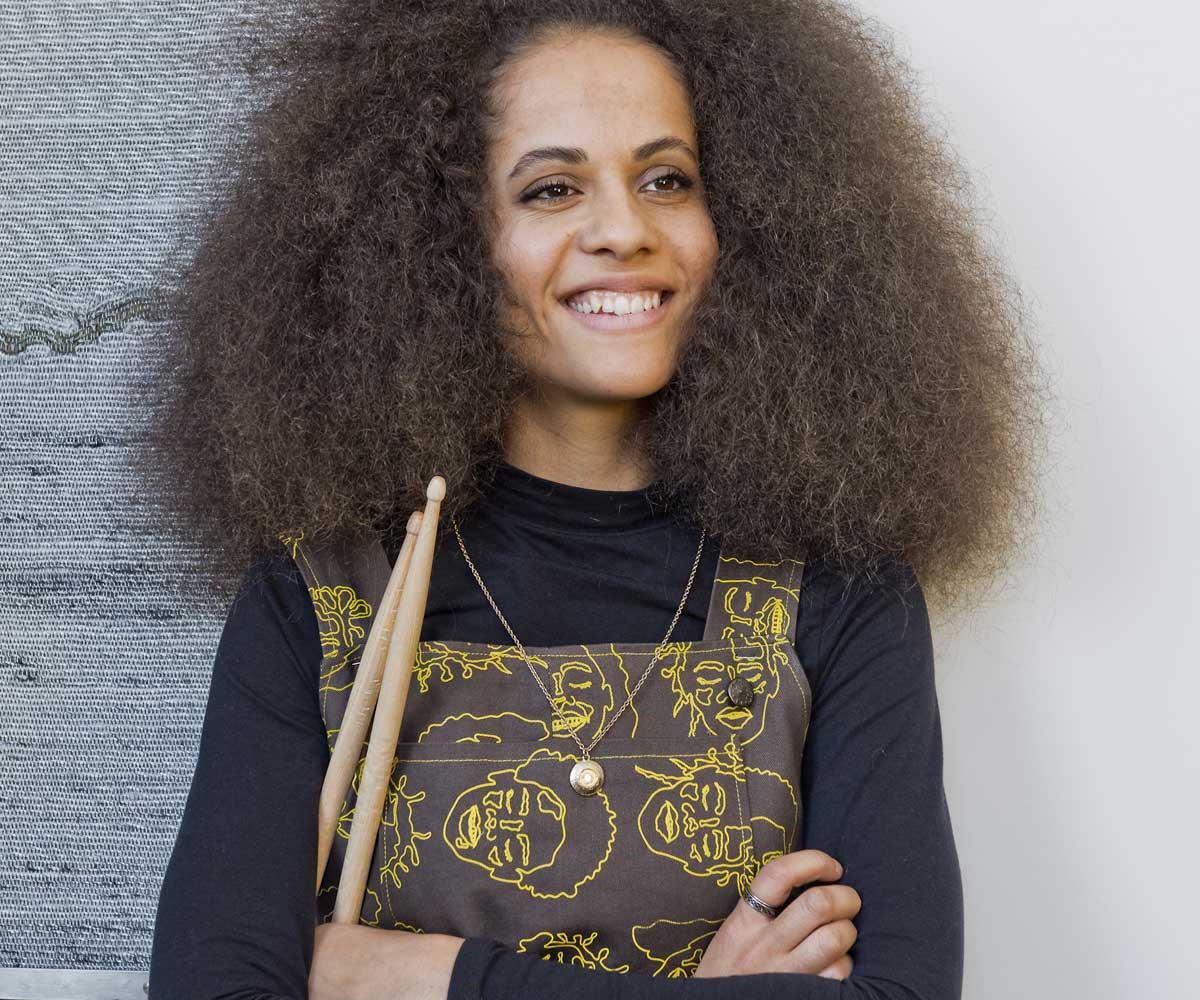 Portrait photograph of drummer Jas Keyser. She is holding a set of drumsticks with arms folded over her chest. She is smiling and confident.