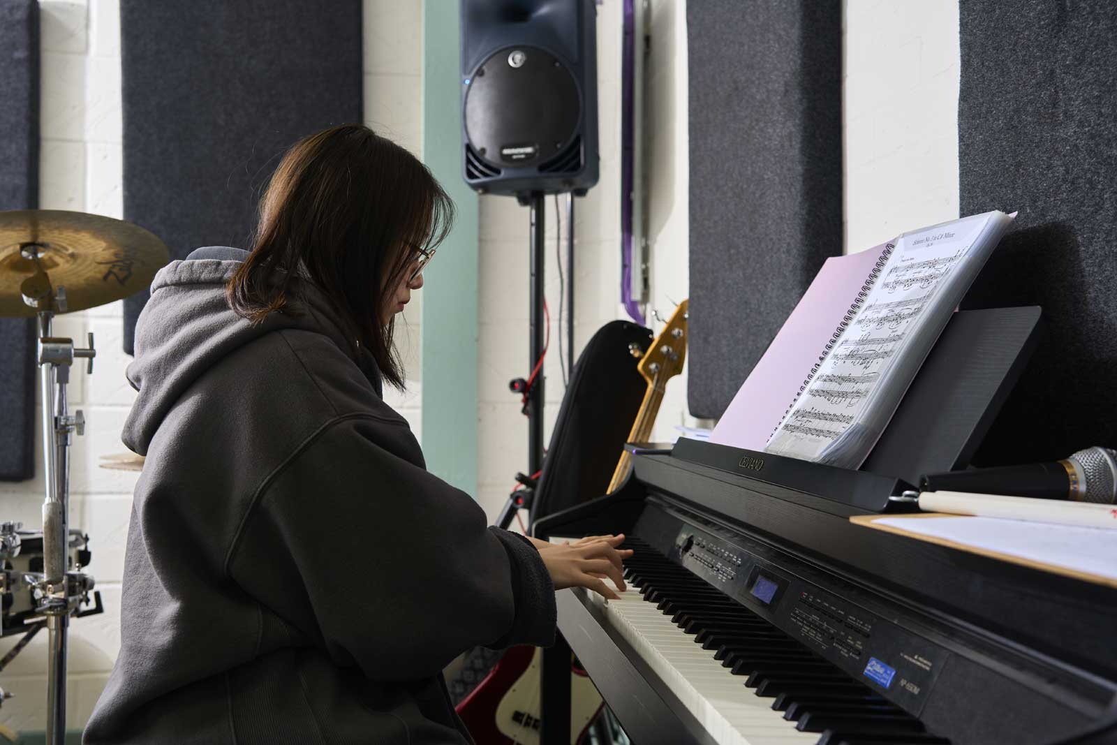 A student sits at a piano in a studio, side on. They are wearing a large hoody.