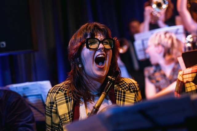 Bess Shooter Close up of Bess laughing exuberantly, she wears dark rimmed glasses and a yellow and black plaid coat. There are some blurred people and music on stands in the background.
