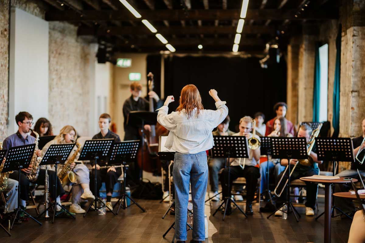 Musical Director Olivia Murphy stands with her back to the camera, she is conducting the Blueprint Orchestra rehearsal.