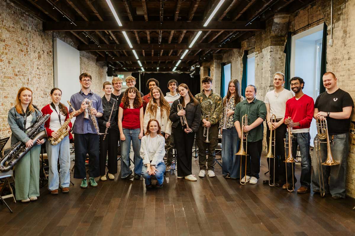 NYJO Blueprint Orchestra stand together looking at the camera. Musical director crouches in front of them in the middle.