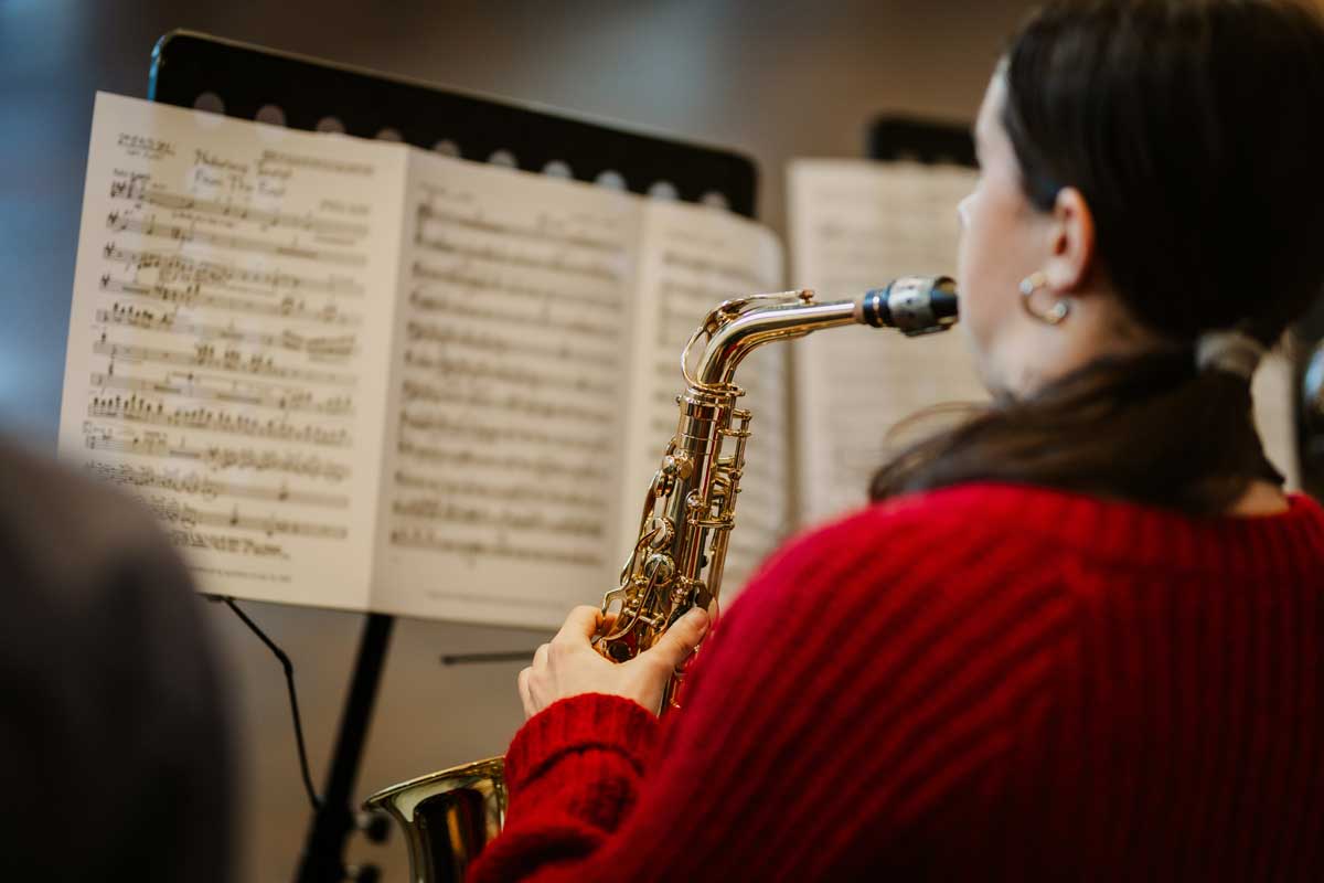 A female saxophonist sits with her back to the camera, playing and reading from sheet music on a music stand. She is wearing a striking red jumper.