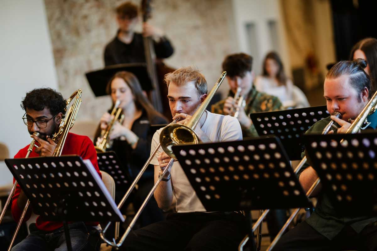 Three trombonists sit behind music stands playing their instruments. 