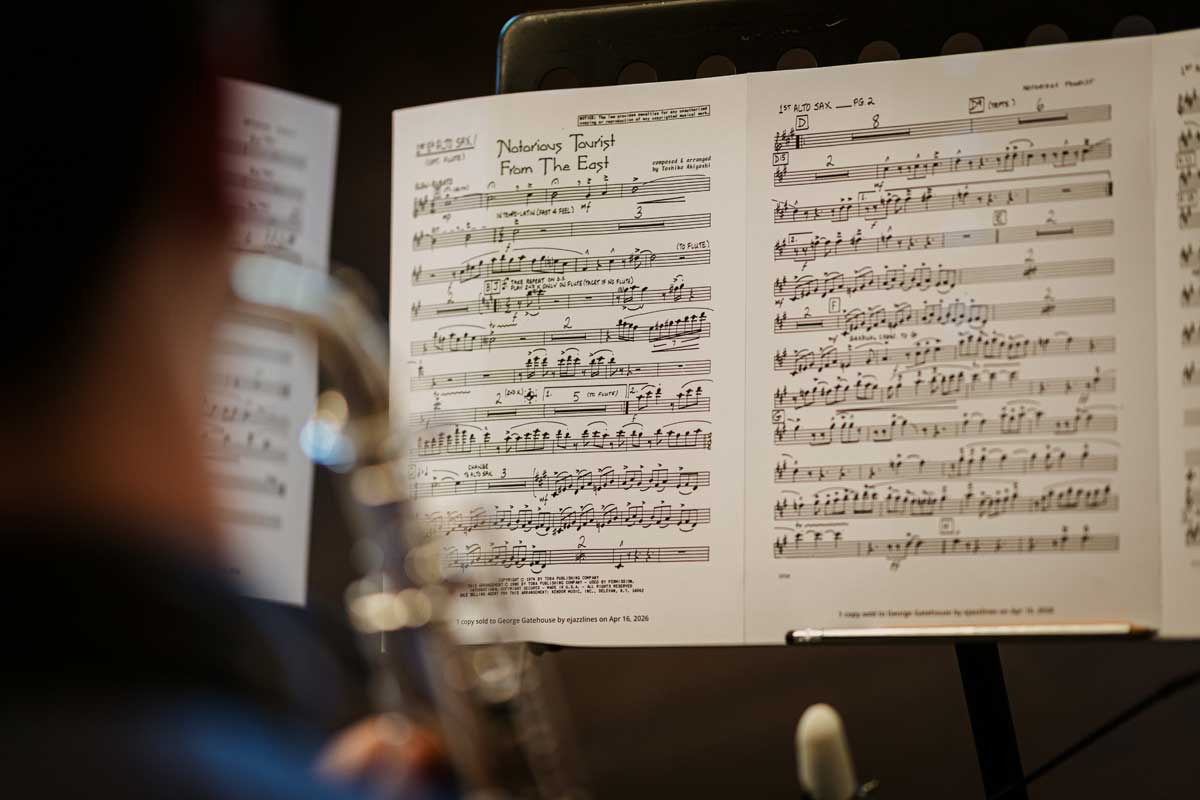 A close up of a jazz chart for the piece 'Notorious Tourist from the East' by Toshiko Akiyoshi. In the foreground is a the head and shoulders of a player with their saxophone, blurred.