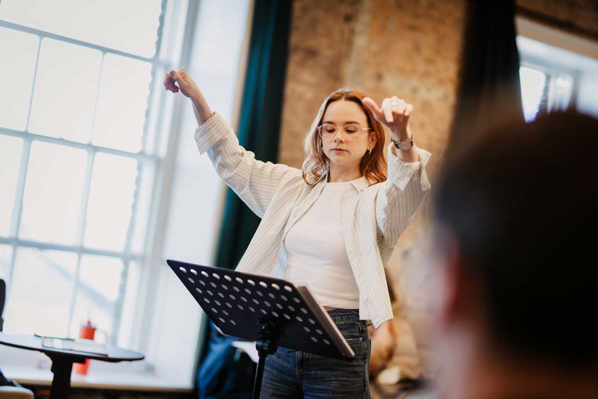 A close up of orchestra MD Olivia Murphy who stands behind a music stand with arms raised. She is wearing a white shirt over a white shirt 