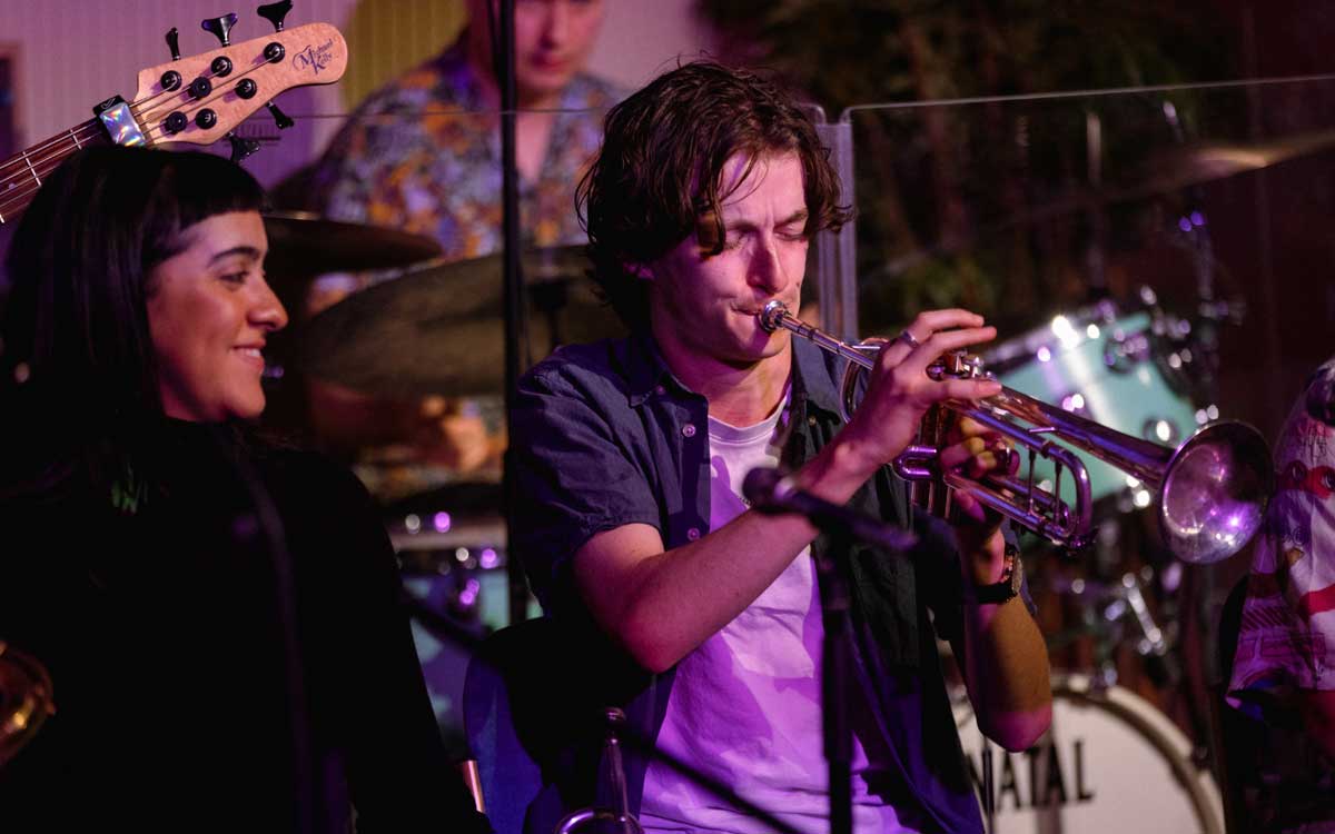 Miles Davis colour 1200x750 Trumpeter Tom Stringer is playing a solo at a gig, he looks relaxed in a white tshirt and open shirt. Band member looks on smiling.
