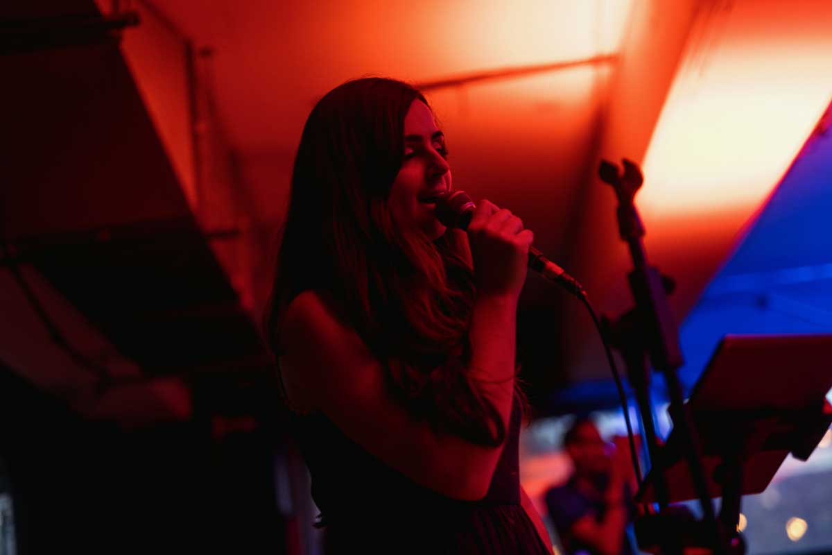 A side on head and shoulders photo of a singer performing, microphone in hand. The background is warm orange lighting with a touch of blue